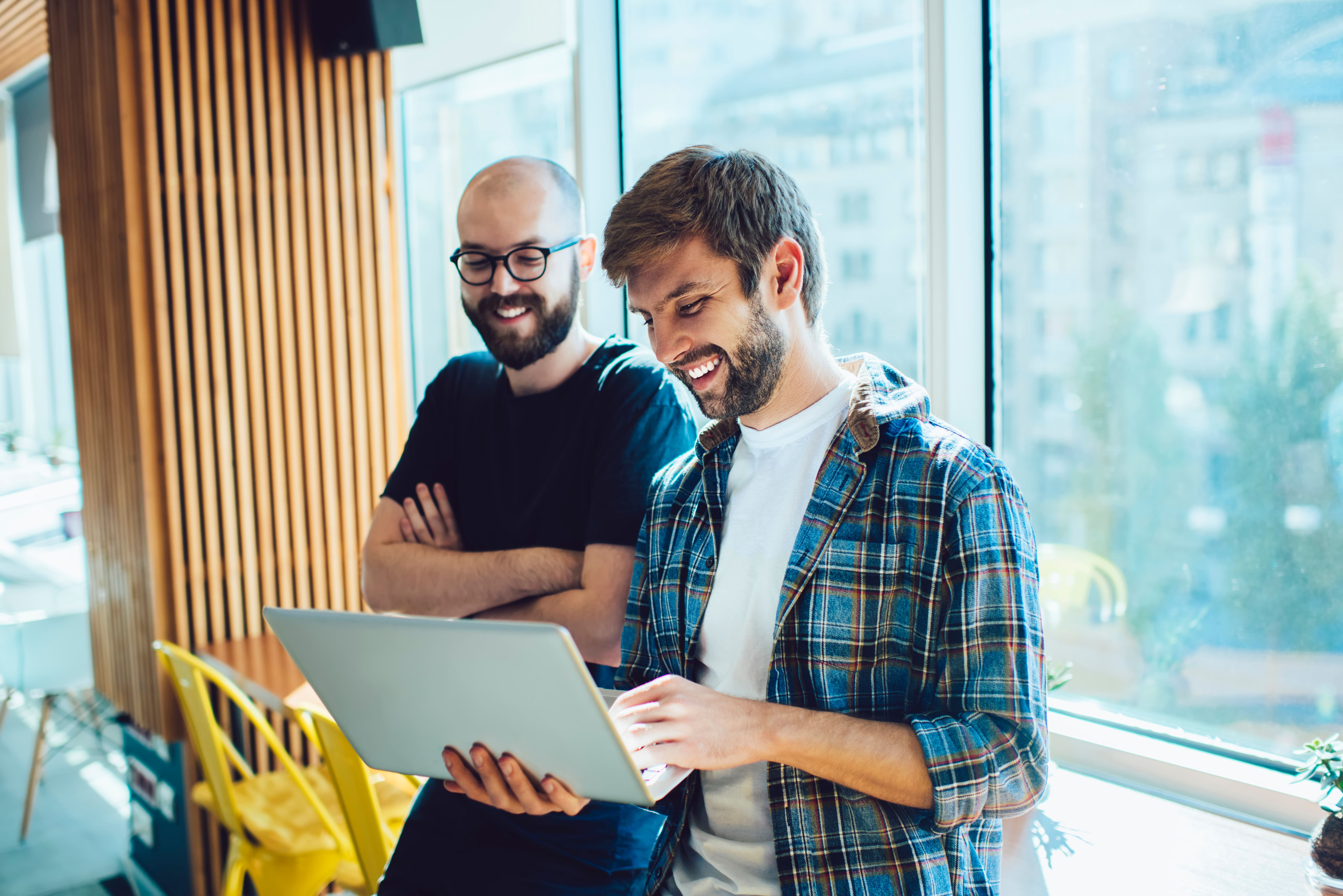 Zwei Männer stehen vor einer Glasfront, der vordere hält einen Laptop in der Hand, beide schauen lachend auf den Bildschirm. 